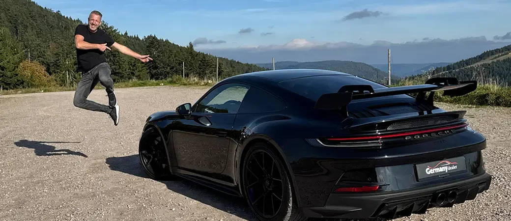 Springend van plezier naast een zwarte Porsche 911 GT3 met Germany Dealer-kentekenplaat